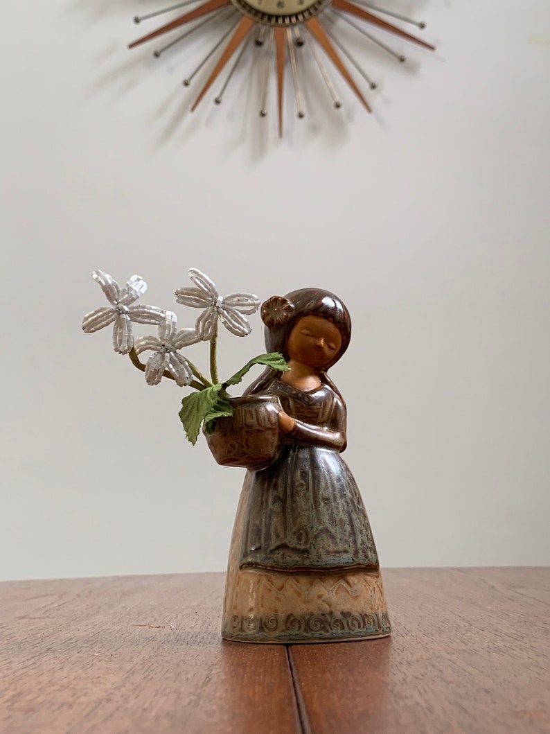 May include: A ceramic figurine of a girl holding a brown pot with white flowers. The girl is wearing a brown dress with a green and brown pattern. The figurine is on a wooden table.