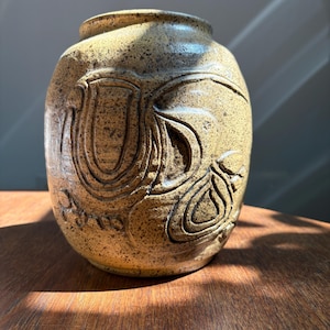 May include: A handcrafted, speckled ceramic vase with an oval shape. The vase features a carved, repeating floral design in a warm, earthy tone. The vase is sitting on a wooden surface.