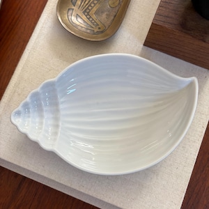 May include: A white ceramic dish shaped like a seashell with a ribbed texture and a pointed end. The dish has a smooth, glossy finish and a curved, open interior. It is positioned next to a small, oval-shaped brass tray with etched details.
