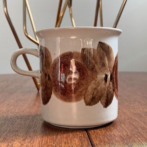 Peut inclure: Une tasse en céramique blanche avec un motif floral marron. La tasse a une anse incurvée et un bord légèrement évasé. Le motif floral présente de grandes fleurs marron stylisées sur un fond blanc. La tasse est posée sur une surface en bois.