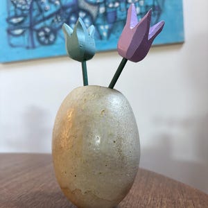May include: A small, egg-shaped vase with a textured, off-white finish. Two wooden tulips, one light blue and one lavender, are inserted into the vase. The stems are green. A blue abstract painting hangs in the background.