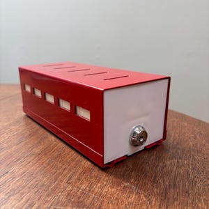 May include: A rectangular red metal box with a white end. The red top has four parallel slits. The side has six small rectangular windows. A silver lock is on the white end. The box is on a wooden surface.
