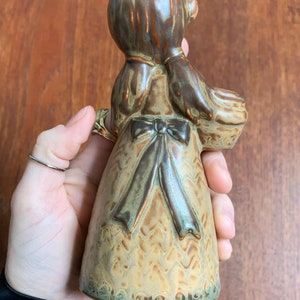 May include: A ceramic figurine of a girl wearing a brown dress with a green and brown bow. The figurine is holding a basket in her right hand.