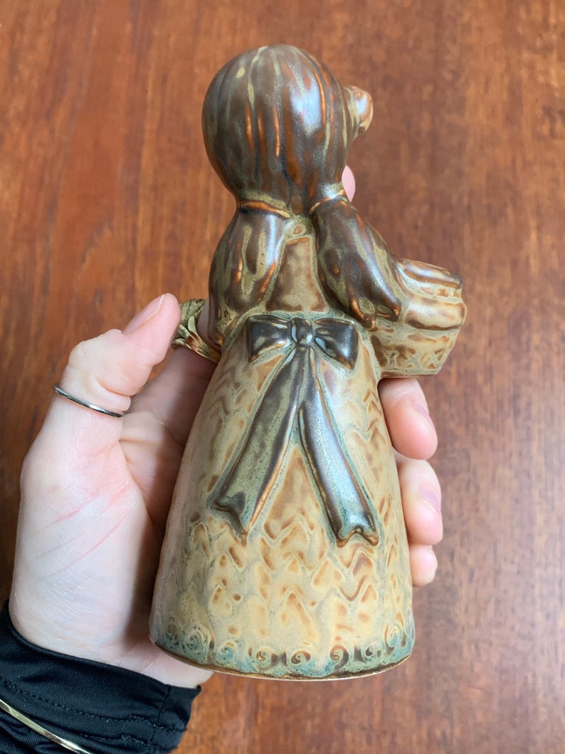 May include: A ceramic figurine of a girl wearing a brown dress with a green and brown bow. The figurine is holding a basket in her right hand.