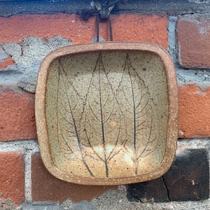 Puede incluir: Un plato cuadrado de cerámica con un esmalte marrón y un diseño de hojas. El plato está colgado en una pared de ladrillo con un cordón marrón.