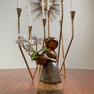 May include: A ceramic figurine of a girl holding a basket of white flowers. The figurine is brown and green with a textured finish. The figurine is standing on a wooden table in front of a gold candelabra.