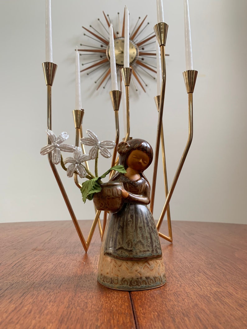 May include: A ceramic figurine of a girl holding a basket of white flowers. The figurine is brown and green with a textured finish. The figurine is standing on a wooden table in front of a gold candelabra.