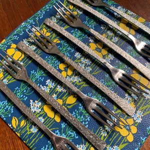 May include: A set of six silver forks with textured handles, arranged on a blue and yellow floral patterned surface.