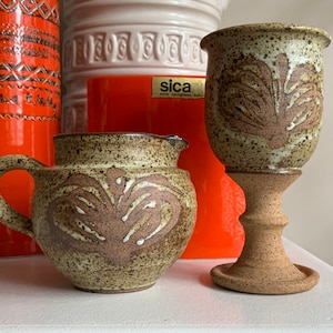 May include: Two ceramic pieces with a speckled brown glaze and a white design. A pitcher with a handle and a goblet with a stem.