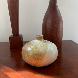 May include: A small, round, ceramic vase with a white and brown glaze. The vase is sitting on a wooden surface.
