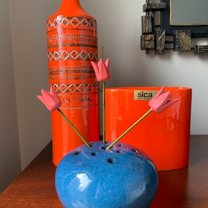 Peut inclure: Un vase en céramique bleu avec une forme ronde et des trous pour les tiges. Le vase est rempli de trois tulipes en bois roses. Le vase est posé sur une table en bois à côté d'un grand vase en céramique orange avec des motifs géométriques noirs et d'un petit vase en céramique orange avec le mot "sica" imprimé dessus.