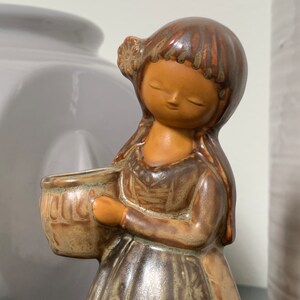 May include: A ceramic figurine of a girl with brown hair and a brown dress holding a brown pot. The figurine has a glazed finish and is decorated with brown and green patterns.
