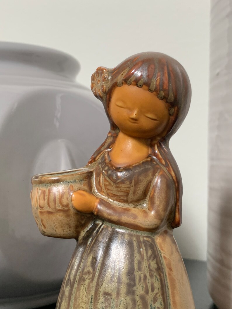 May include: A ceramic figurine of a girl with brown hair and a brown dress holding a brown pot. The figurine has a glazed finish and is decorated with brown and green patterns.
