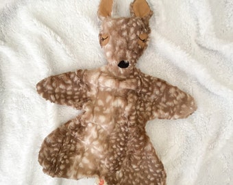 May include: A fawn-shaped baby comforter with a brown and white spotted pattern. The fawn has tan ears, closed eyes, and a black nose. The comforter is on a white, fluffy blanket.