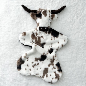 May include: A white and brown plush cow-shaped baby blanket with a black nose and eyes. The blanket is made of soft, fuzzy fabric and is perfect for cuddling.