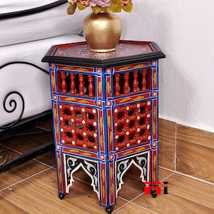 May include: A hand-painted, hexagonal side table with intricate geometric designs. The table features a red, blue, and gold color scheme with a black top and decorative cutouts. A gold vase with flowers sits on top. The table is approximately 24 inches tall.