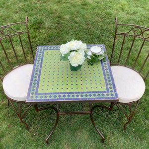 May include: A small, square, green and blue mosaic tile table with a metal frame. Two metal chairs with white cushions surround the table. A bouquet of white flowers sits in the center of the table.