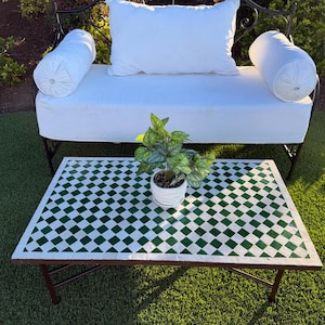 May include: Outdoor scene featuring a white cushioned bench with bolster pillows and a rectangular coffee table. The table has a green and white diamond pattern. A small potted plant sits on the table, adding a touch of nature to the setting.
