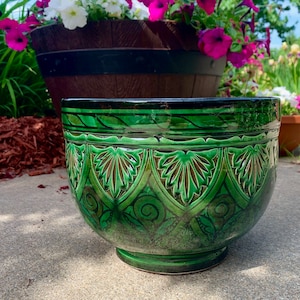 May include: A green ceramic planter with intricate embossed designs. The planter has a wide, round base and a slightly flared rim.