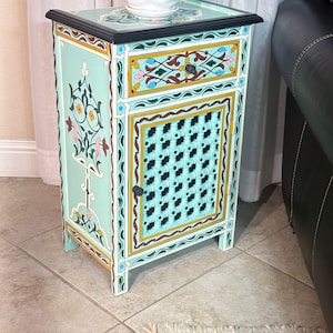 May include: A small, hand-painted wooden nightstand with a single drawer and a cabinet door. The nightstand is painted in a light blue color with intricate floral designs in yellow, pink, and green. The cabinet door has a lattice design and a black knob.