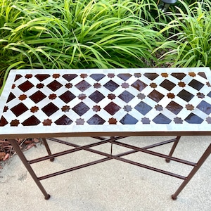 May include: A rectangular outdoor side table with a mosaic tile top. The tabletop features a repeating pattern of brown and white diamonds and star shapes. The table has a dark brown metal frame and legs.