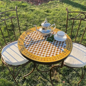 May include: A small bistro set with a mosaic tile tabletop in shades of yellow and brown. The set includes two wrought iron chairs with cream-colored cushions, and a silver teapot and teacup. The set is outdoors on a grassy lawn.