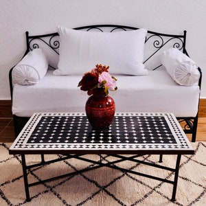 May include: A black metal daybed with white cushions and a black and white mosaic tile top coffee table. A bouquet of pink and red flowers in a red vase sits on the table.