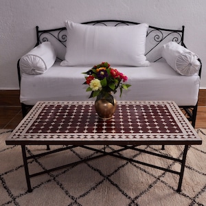 May include: A dark red and white tiled coffee table with a black metal frame. The table is sitting on a white and black patterned rug in front of a white couch with black metal accents.