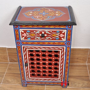 May include: A small, hand-painted wooden nightstand with a red base, blue and yellow accents, and intricate floral designs. It features a drawer and a cabinet door with a lattice design.