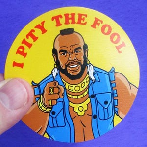 May include: Round yellow button with the phrase "I PITY THE FOOL" in red lettering. The button features a cartoon illustration of a man with a mohawk, gold jewelry, and a blue denim vest, pointing at the viewer.