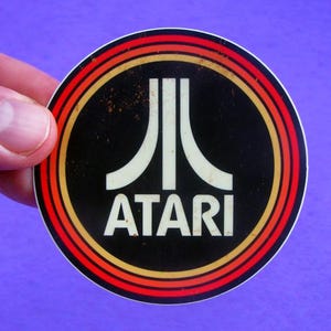 May include: A round Atari sticker with a black background and a white Atari logo. The logo is surrounded by concentric rings of red, orange, and yellow. The word "ATARI" is printed in white below the logo.