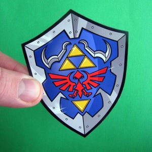 May include: A blue shield sticker with a silver border and rivets. The shield features a red bird emblem, three yellow triangles, and silver crescent shapes. The shield is held against a green background.