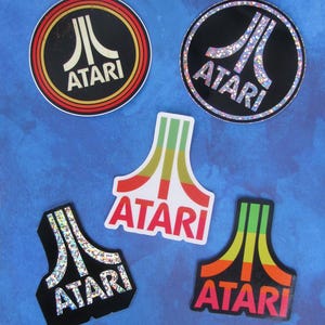 Può includere: Una collezione di adesivi Atari in varie forme e design. Gli adesivi presentano il logo Atari in diverse combinazioni di colori, tra cui nero, rosso e accenti glitterati. Gli adesivi sono su uno sfondo blu.