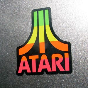 Może przedstawiać: Kolorowa naklejka z logo Atari. Naklejka ma czarne tło ze słowem "ATARI" w różowych literach. Nad słowem znajdują się pionowe paski w kolorze zielonym, żółtym i pomarańczowym. Naklejka ma czarną obwódkę.