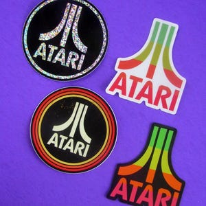 Puede incluir: Cuatro pegatinas con el logotipo de Atari en varios diseños. Dos pegatinas circulares con el logotipo y el texto de Atari, una con fondo negro y detalles brillantes, la otra con fondo negro y anillos rojos y amarillos. Dos pegatinas con el logotipo de Atari en un diseño estilizado.