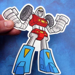 May include: A sticker featuring a cartoon robot with a red torso, gray head, and blue legs. The robot has gray arms with tires for hands. Yellow accents are on the robot's chest and legs. The sticker is held by a hand.