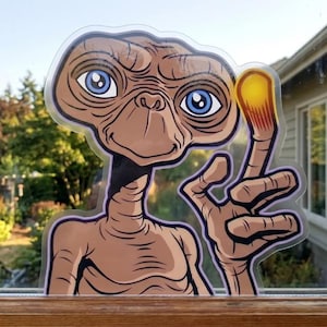 May include: A window cling featuring the character E.T. The alien has large blue eyes, a tan complexion, and a glowing finger. The cling is placed on a window with a view of trees and a house.