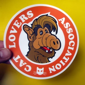 May include: A circular sticker with an orange border featuring the text CAT LOVERS ASSOCIATION and a small cat icon. In the centre, a brown, furry, puppet-like creature with a large nose, dark hair, and an open mouth is depicted on a white background.