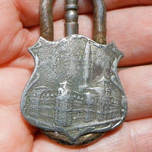 May include: A vintage silver-toned metal souvenir padlock. The padlock features a shield-shaped face with an embossed cityscape and a tall building. The lock's handle is a curved design. The metal has a worn, aged appearance.