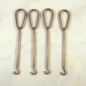 May include: Four silver-colored metal crochet hooks are arranged horizontally. Each hook has a teardrop-shaped handle and a curved hook at the bottom. The hooks are evenly spaced against a white background.