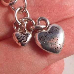 Peut inclure: Gros plan sur un bracelet de charme argent&eacute; avec deux breloques en forme de c&oelig;ur. L'un des charms porte la lettre "B" grav&eacute;e, et l'autre le mot "Kenya". Les charms sont reli&eacute;s par des anneaux argent&eacute;s.