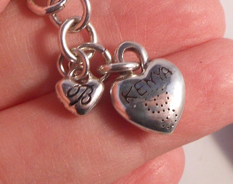 Peut inclure: Gros plan sur un bracelet de charme argent&eacute; avec deux breloques en forme de c&oelig;ur. L'un des charms porte la lettre "B" grav&eacute;e, et l'autre le mot "Kenya". Les charms sont reli&eacute;s par des anneaux argent&eacute;s.