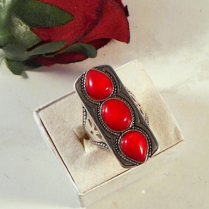 May include: A silver-toned ring featuring three vibrant red oval-shaped stones. The ring has an elongated rectangular design with intricate detailing around the stones and on the band. The ring is presented in a white jewelry box.