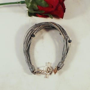 May include: A multi-strand bracelet composed of thin, metallic silver-toned wires gathered at each end by coiled silver-toned metal bands. The bracelet features a silver-toned toggle clasp with a hexagonal ring and a small, dangling triangular charm.