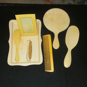 Può includere: Un set da toeletta antico nei toni crema e giallo. Il set comprende un vassoio, una piccola cornice, un pettine, uno specchio a mano e una spazzola per capelli. Gli oggetti sono disposti su una superficie scura.