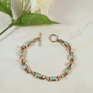 May include: A gold-toned bracelet featuring rectangular turquoise-colored stones. The bracelet has a toggle clasp and is displayed on a white surface with a flower in the background. The bracelet is a delicate piece of jewelry.