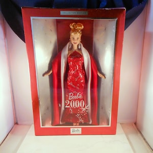 May include: A Barbie Collector Edition doll in a red box. The doll is wearing a red sequined gown and a white and red cape. The box has the text "Barbie 2000" and "Collector Edition".