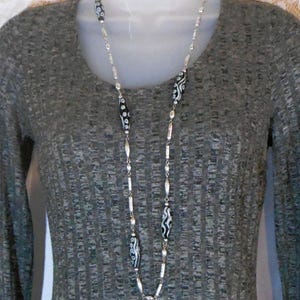Peut inclure: Un long collier argent&eacute; avec des perles &agrave; motifs noirs et blancs. Le collier est compos&eacute; d'une cha&icirc;ne d&eacute;licate parsem&eacute;e de petites perles rondes. Les perles noires et blanches pr&eacute;sentent un motif abstrait unique. Le collier est pr&eacute;sent&eacute; sur un fond gris.