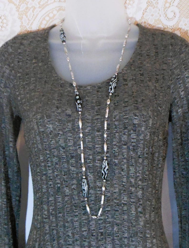 Peut inclure: Un long collier argent&eacute; avec des perles &agrave; motifs noirs et blancs. Le collier est compos&eacute; d'une cha&icirc;ne d&eacute;licate parsem&eacute;e de petites perles rondes. Les perles noires et blanches pr&eacute;sentent un motif abstrait unique. Le collier est pr&eacute;sent&eacute; sur un fond gris.