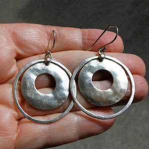 Peut inclure: Une paire de boucles d'oreilles argentées au design circulaire, composées d'un disque texturé plus petit suspendu à l'intérieur d'un anneau plus grand. Elles sont fixées à des crochets incurvés et présentent une finition métallique nuancée.